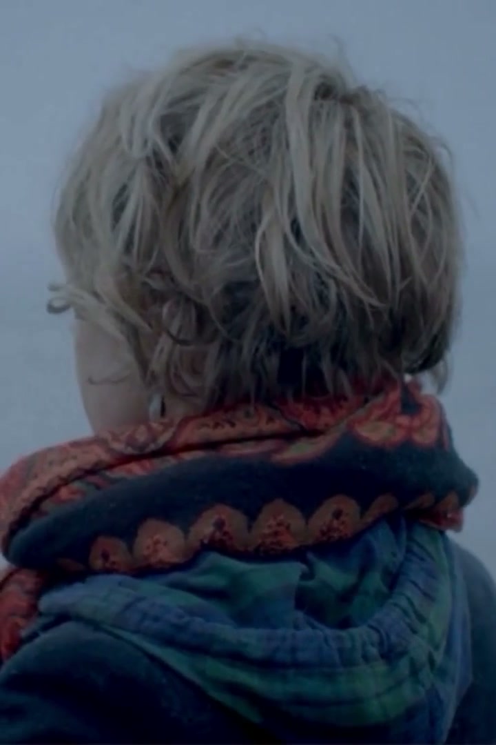 A child with tousled fair hair and a red patterned scarf, seen from behind, looks out over a grey sea